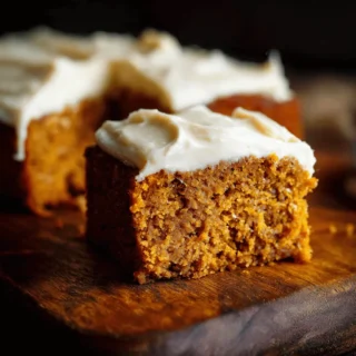 A close-up of a frosted slice from a delicious homemade pumpkin cake recipe on a dark wooden surface.
